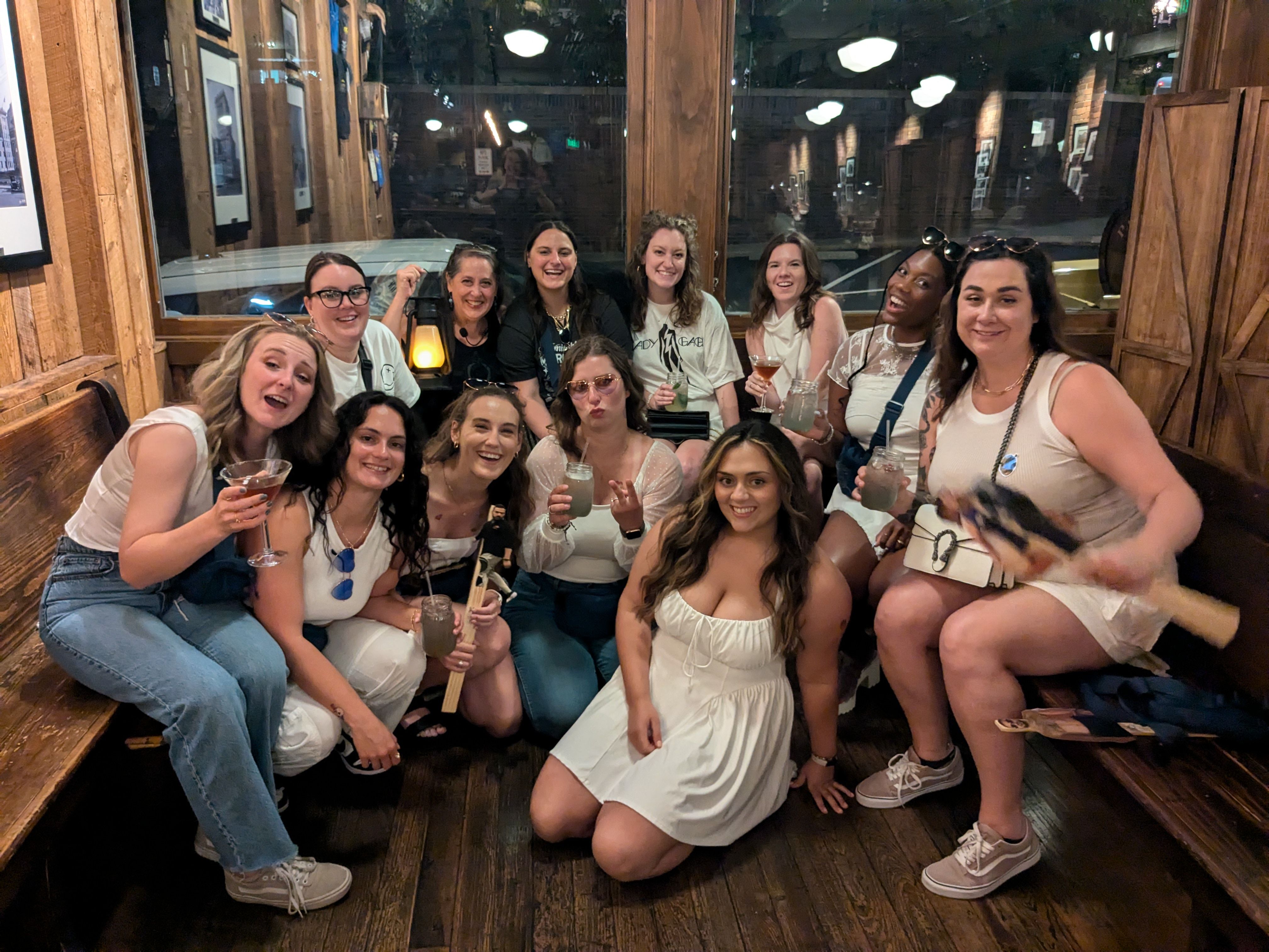 Group of friends in a rustic wood-paneled bar, smiling and holding cocktails for a lively girls' night out with warm ambient lighting and bench seating.