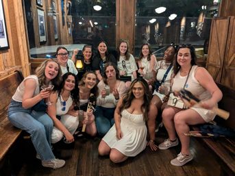 Group of friends in a rustic wood-paneled bar, smiling and holding cocktails for a lively girls' night out with warm ambient lighting and bench seating.