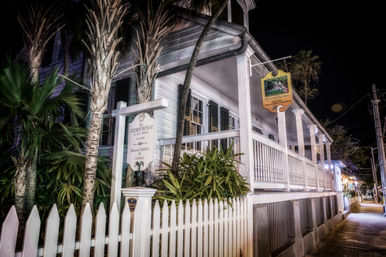 Charming historic white wooden house with wraparound porch, white picket fence and palm trees glowing under night street lights