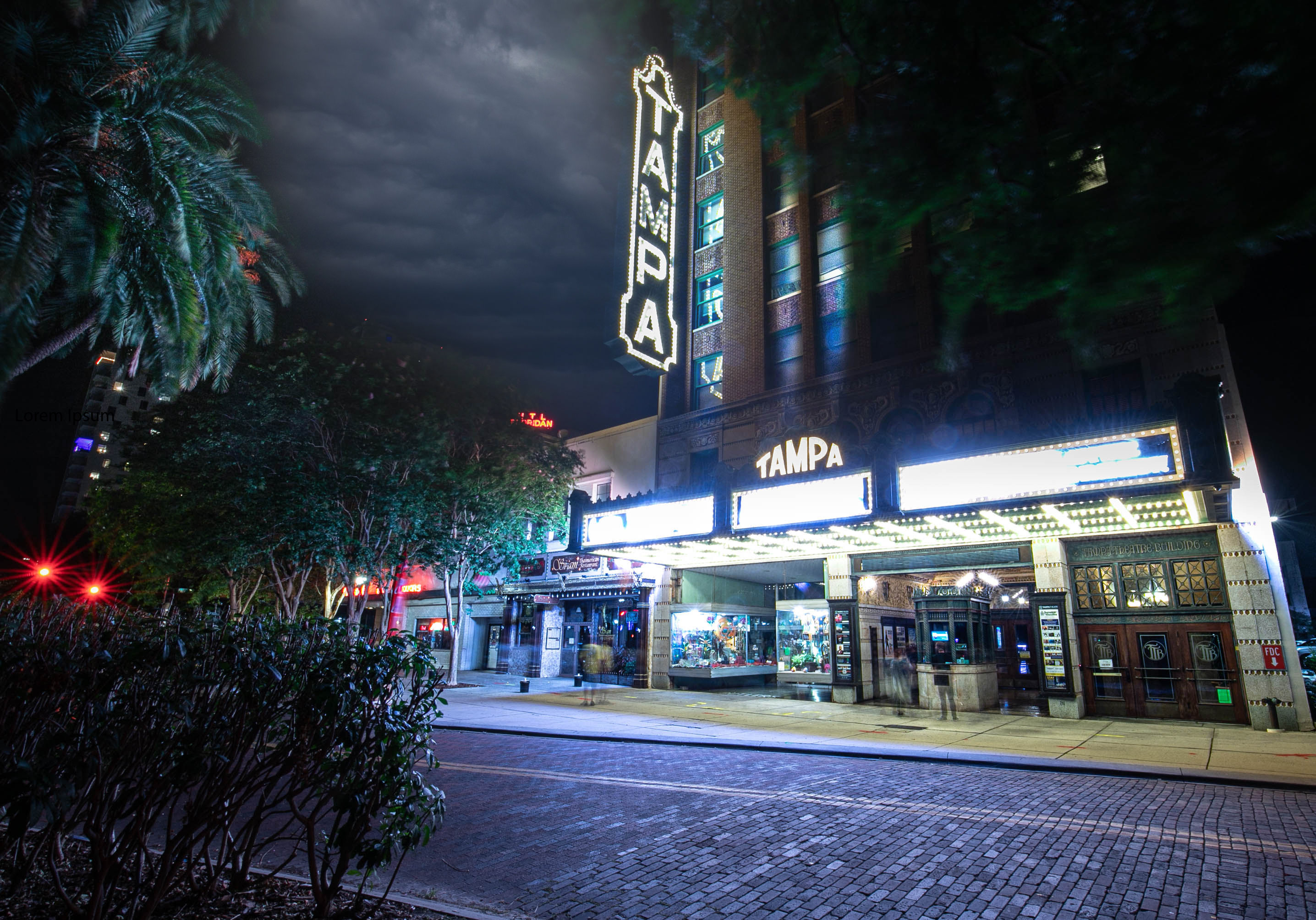 Nighttime scene of a historic Tampa theater with a glowing vertical "TAMPA" neon sign and illuminated marquee, palm trees, empty brick street and moody cloudy sky.