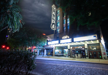 Nighttime scene of a historic Tampa theater with a glowing vertical "TAMPA" neon sign and illuminated marquee, palm trees, empty brick street and moody cloudy sky.