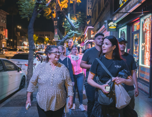 Group of people walking along a neon-lit downtown sidewalk at night, chatting past tattoo shop signs, parked cars, trees and busy traffic
