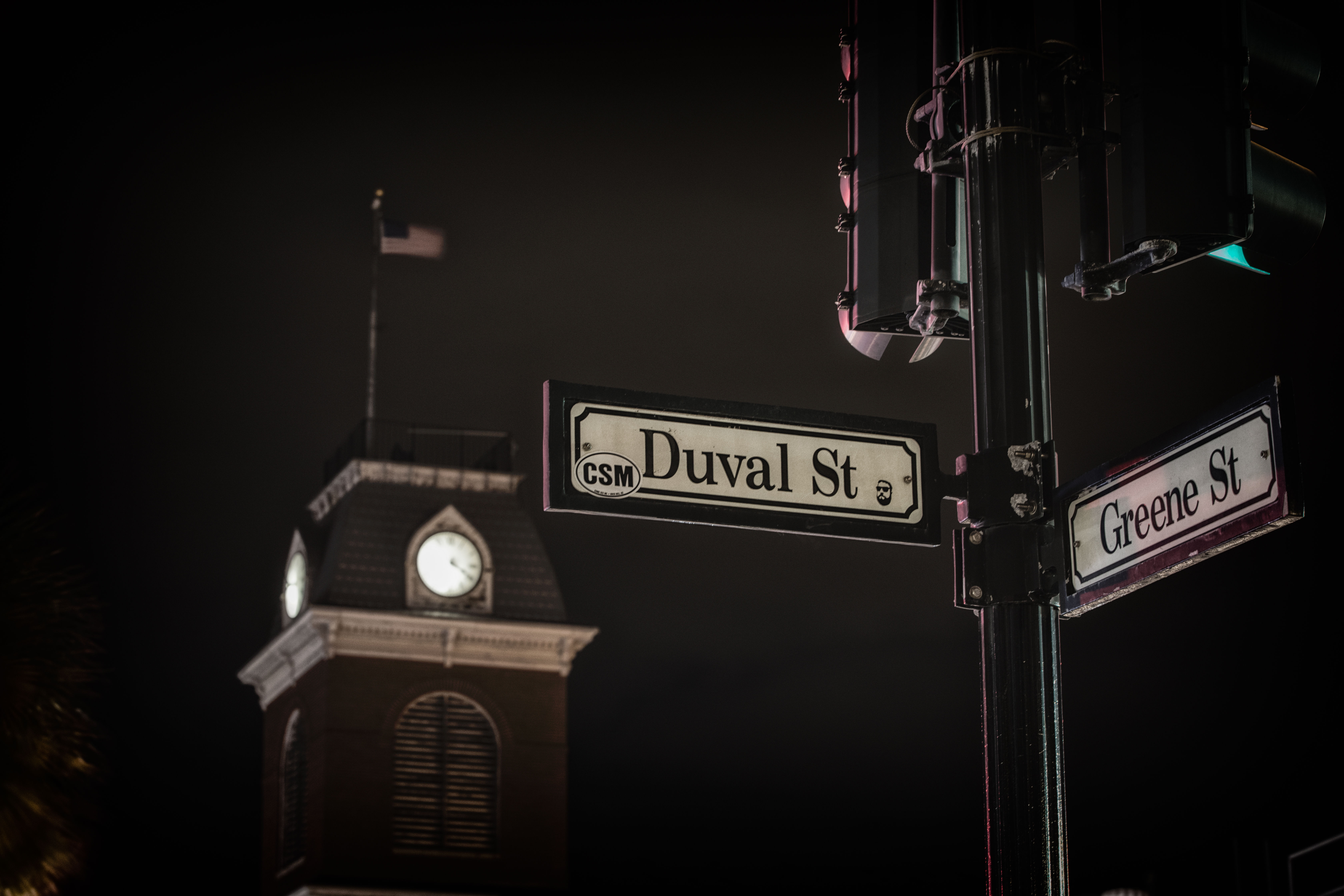 Moody nighttime scene of Duval St and Greene St signs on a lamppost, with an illuminated clock tower and waving flag blurred in the background.