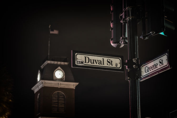 Moody nighttime scene of Duval St and Greene St signs on a lamppost, with an illuminated clock tower and waving flag blurred in the background.