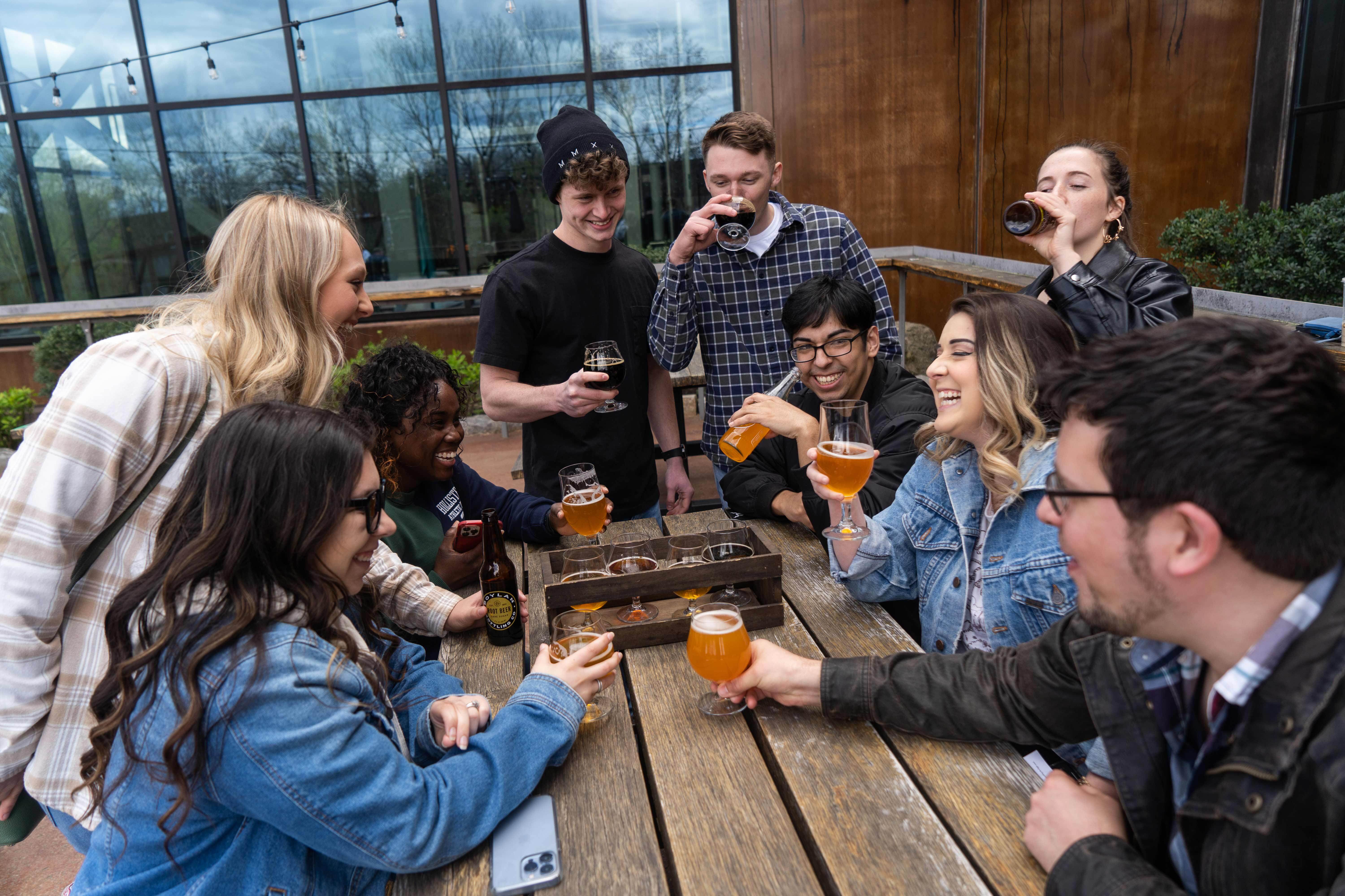 Group of friends laughing and enjoying craft beers and a flight on a wooden picnic table at an outdoor brewery patio with glass windows and string lights.