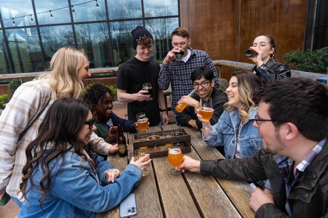 Group of friends laughing and enjoying craft beers and a flight on a wooden picnic table at an outdoor brewery patio with glass windows and string lights.