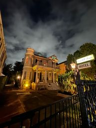 Historic Victorian-style hotel exterior at night — turret and columned porch bathed in warm lights, ornate iron fence and glowing streetlamp in the foreground, dramatic cloudy sky overhead.