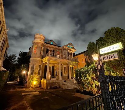 Historic Victorian-style hotel exterior at night — turret and columned porch bathed in warm lights, ornate iron fence and glowing streetlamp in the foreground, dramatic cloudy sky overhead.