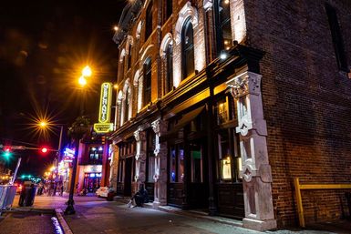 Vibrant downtown nightlife scene with a historic brick building featuring illuminated arched windows, ornate stone columns and a restaurant entrance, neon signs and glowing streetlights on a lively urban block.