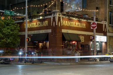 Nighttime urban nightlife scene: a brick corner bar with rooftop patio and string lights, a neon bar sign, parked scooters and streaking car light trails.