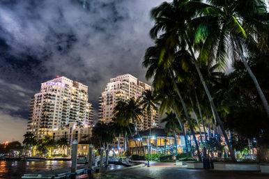 Tropical waterfront at night with illuminated high-rise condos, palm-lined marina promenade and dramatic cloudy sky