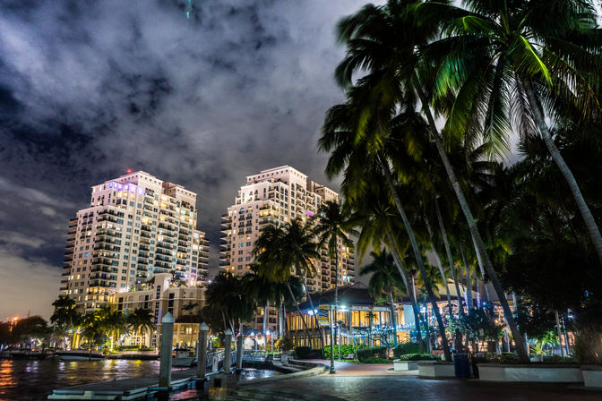Tropical waterfront at night with illuminated high-rise condos, palm-lined marina promenade and dramatic cloudy sky