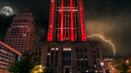 Downtown night scene featuring an art-deco skyscraper glowing red among high-rises, dramatic lightning bolt and full moon over an illuminated city street.