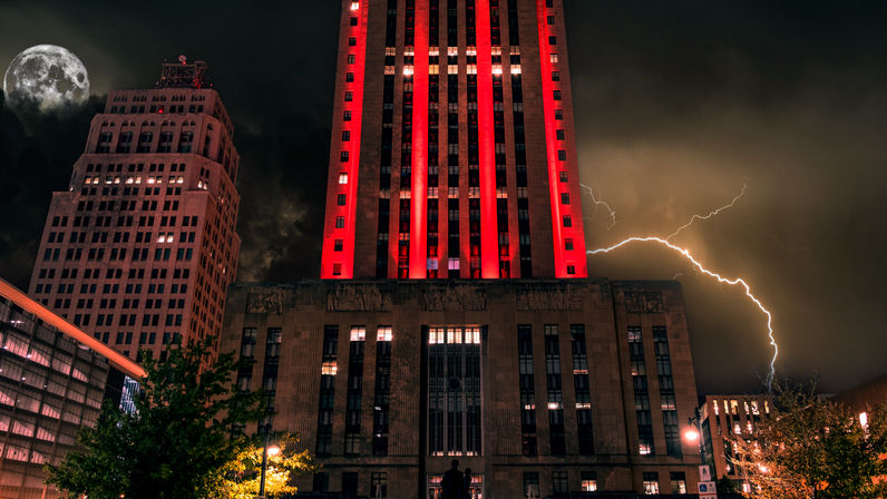 Downtown night scene featuring an art-deco skyscraper glowing red among high-rises, dramatic lightning bolt and full moon over an illuminated city street.