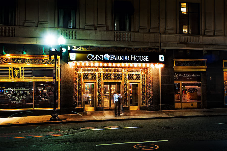 Night view of a historic downtown hotel entrance in Boston with ornate gold-trimmed marquee lights, revolving doors, a streetlamp, and two blurred pedestrians on the sidewalk.
