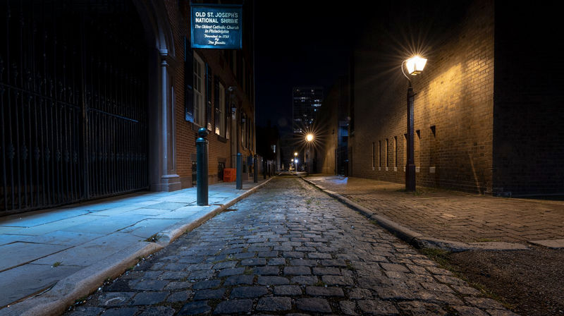 Moody, lamp-lit night view of a historic cobblestone alley lined with brick buildings and a blue-lit sidewalk, city lights visible in the distance