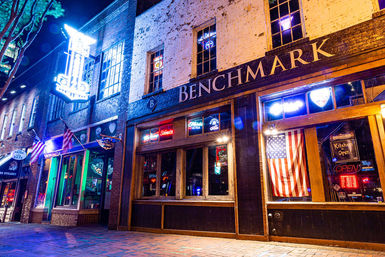 Vibrant nighttime downtown barfront with brick facade, glowing neon signs, an American flag in the window, and an empty brick sidewalk.