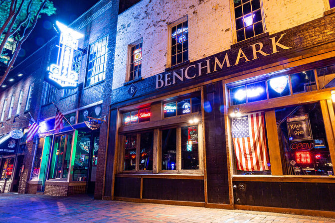 Vibrant nighttime downtown barfront with brick facade, glowing neon signs, an American flag in the window, and an empty brick sidewalk.