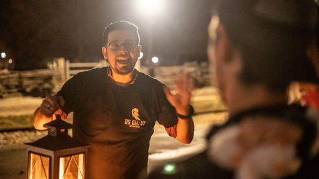 Lantern-lit guide animatedly speaking during a nighttime outdoor ghost tour, warm glow on his face with a wooden fence and dark surroundings in the background.
