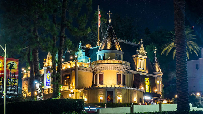 Castle-style theater building lit at night with conical turrets, glowing windows, marquee banners, palm trees and decorative street lamps.