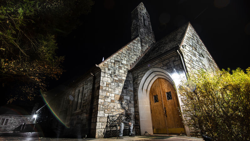 Moody night shot of a historic stone church with a tall steeple and illuminated arched wooden doors, wet sidewalk reflecting light, a person on a bench and trimmed bushes by the entrance.