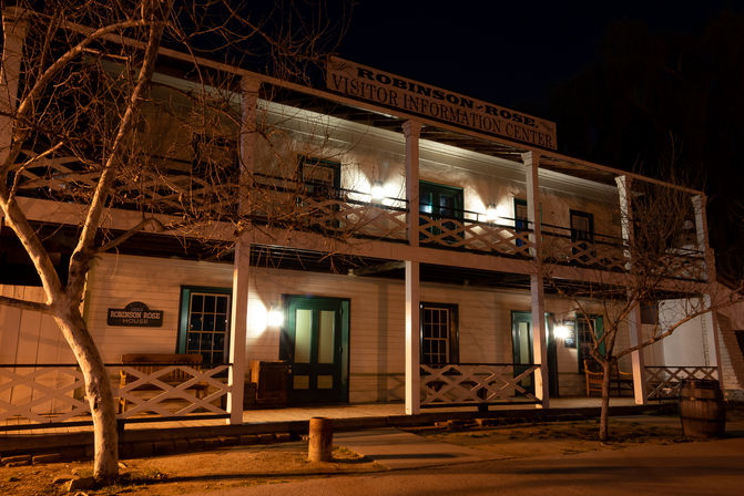 Historic two-story wooden building with a balcony and front porch, warm exterior lights glowing on a quiet small-town street at night