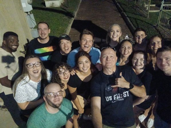 Cheerful nighttime group selfie of about fifteen adults on a brick pathway outdoors, smiling and posing closely around the person holding the camera.