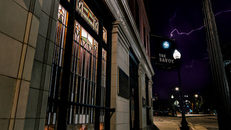 Historic downtown restaurant exterior at night with illuminated stained-glass windows, ornate stone entrance, a glowing streetlamp, and lightning-streaked purple sky over a quiet city intersection.