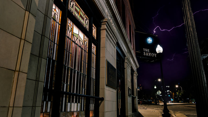 Historic downtown restaurant exterior at night with illuminated stained-glass windows, ornate stone entrance, a glowing streetlamp, and lightning-streaked purple sky over a quiet city intersection.