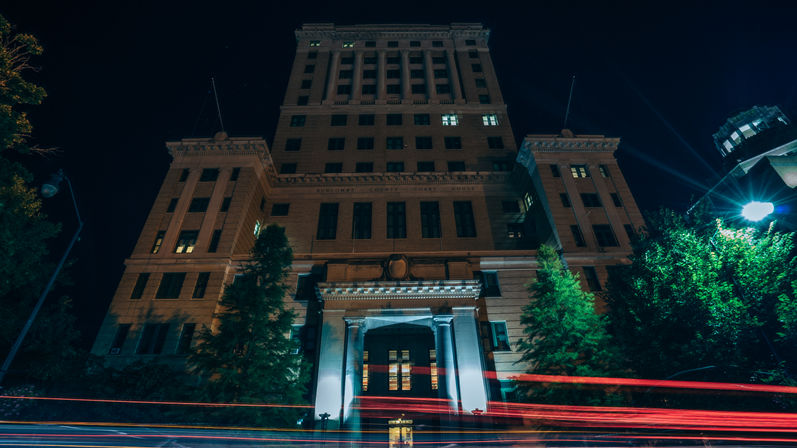 Nighttime view of a historic downtown stone office building with an illuminated columned entrance, tall windows, trees at the facade, and red light trails from passing cars.