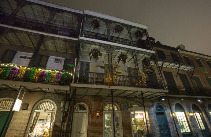 Nighttime French Quarter streetscape with historic brick building and ornate wrought-iron balconies draped in Mardi Gras purple, green and gold beads and string lights, hanging plants and lit shop windows below.