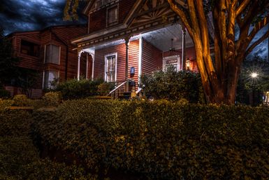 Nighttime view of a historic red wooden Victorian house with a warmly lit front porch, steps and lantern, manicured hedges and a large tree under a dramatic cloudy sky.