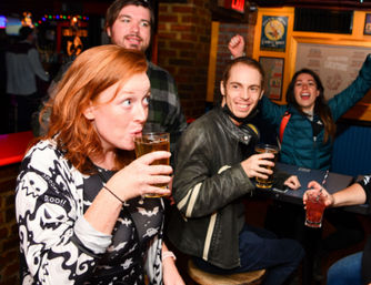 Group of friends enjoying drinks in a lively neighborhood bar — redheaded woman sipping a pint, smiling man holding a beer, and others cheering at a cozy pub high-top table.