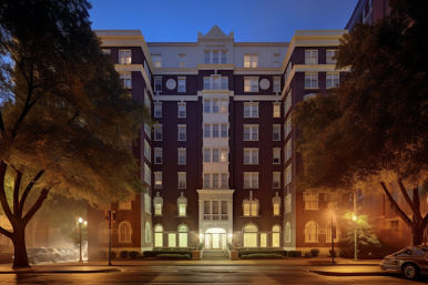 Multi-story historic brick apartment building at dusk on a tree-lined urban street, glowing ornate entrance and street lamps with parked cars nearby.