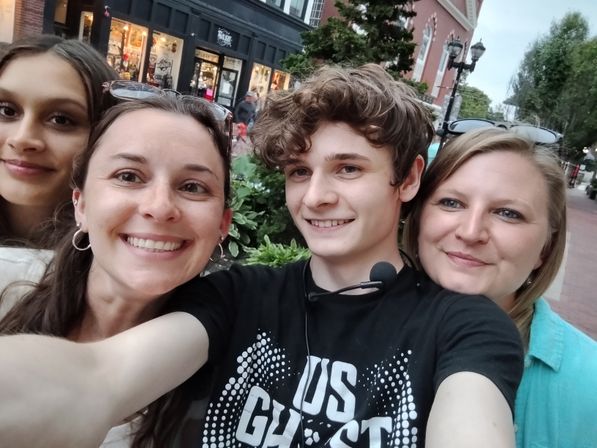 Four smiling people taking a selfie on a downtown brick-lined shopping street with lit storefronts, street lamps, and greenery.