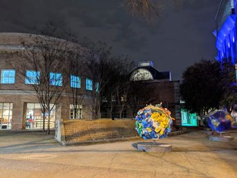 Nighttime urban plaza with a curved brick public building and lit windows, leafless trees, and two illuminated globe sculptures — a colorful recycled-plastic globe and a painted Earth — under blue architectural lighting.