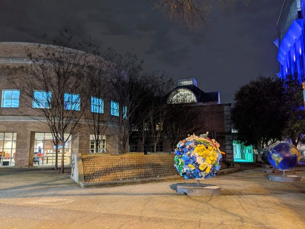 Nighttime urban plaza with a curved brick public building and lit windows, leafless trees, and two illuminated globe sculptures — a colorful recycled-plastic globe and a painted Earth — under blue architectural lighting.