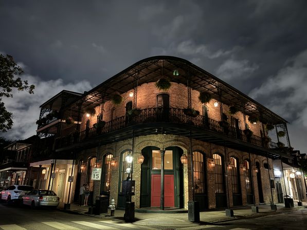 Nighttime New Orleans French Quarter corner building — two-story brick with ornate wrought-iron balcony, hanging plants, warm streetlamps and red double doors under a moody cloudy sky.