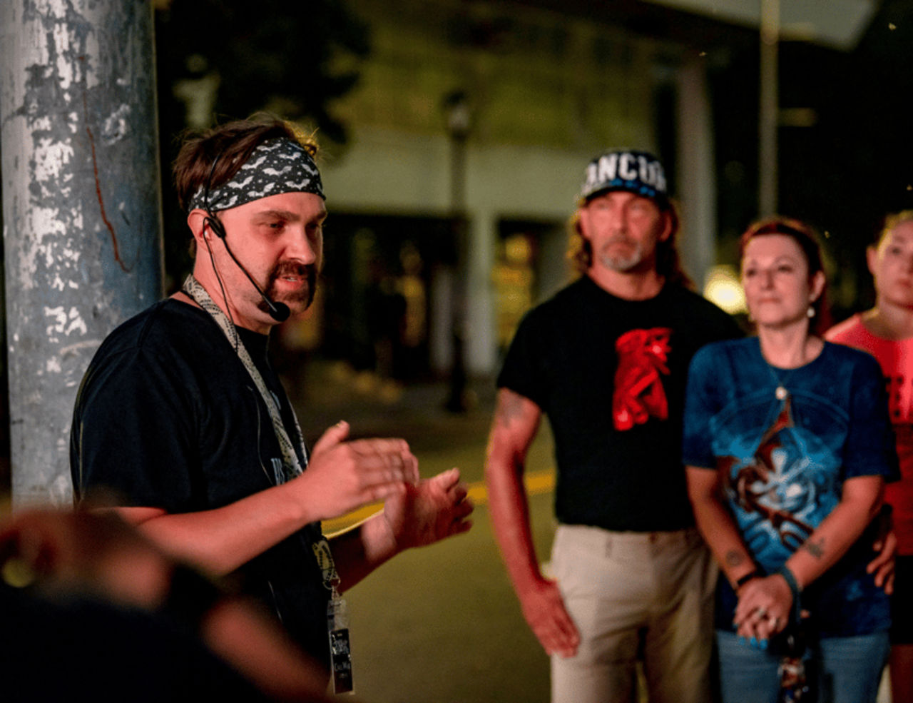 Man wearing a bandana and headset microphone gestures as a small group listens on a dimly lit downtown sidewalk at night