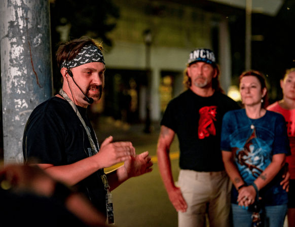 Man wearing a bandana and headset microphone gestures as a small group listens on a dimly lit downtown sidewalk at night