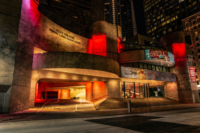 Sleek curved concrete theater entrance in a downtown night scene, glowing with dramatic red and purple accent lights, ramps and steps leading to glass doors under illuminated marquee banners and high-rise buildings.