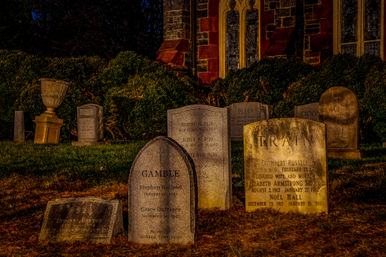 Moody nighttime churchyard with weathered gravestones and stone monuments bathed in warm light in front of a historic stone church with arched windows