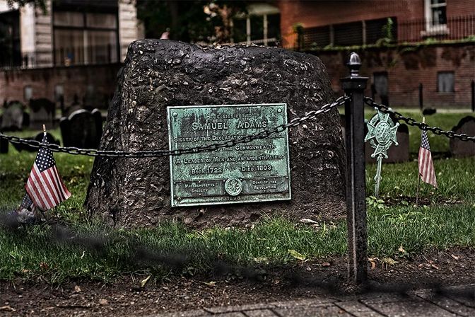 Patinated bronze plaque mounted on a weathered fieldstone monument in a historic cemetery, surrounded by a low chain fence and small American flags.