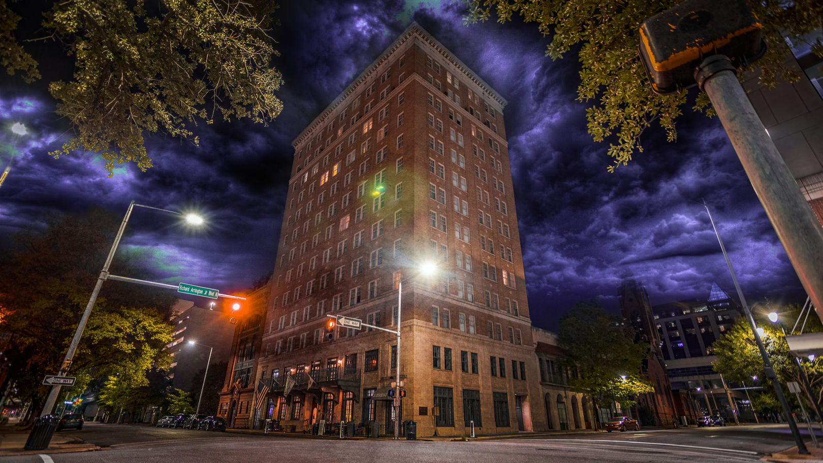 Towering historic brick high-rise on a lit downtown intersection at night, glowing streetlights and traffic signals beneath dramatic purple storm clouds and urban trees.