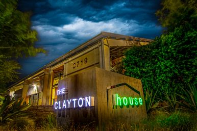 Nighttime view of a modern, neon-lit event venue entrance with white and green signage on a wooden display, industrial-style building, lush landscaping, and dramatic stormy clouds overhead.