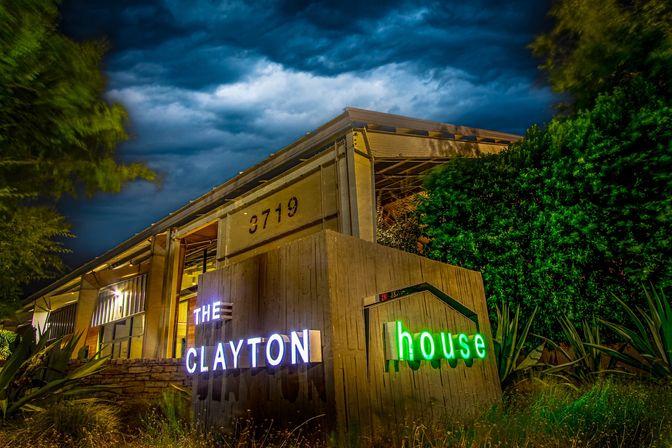 Nighttime view of a modern, neon-lit event venue entrance with white and green signage on a wooden display, industrial-style building, lush landscaping, and dramatic stormy clouds overhead.