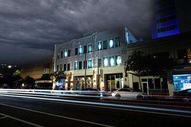 Moody downtown night street with a three-story building glowing blue through windows, storm clouds overhead and streaking white and red car light trails.