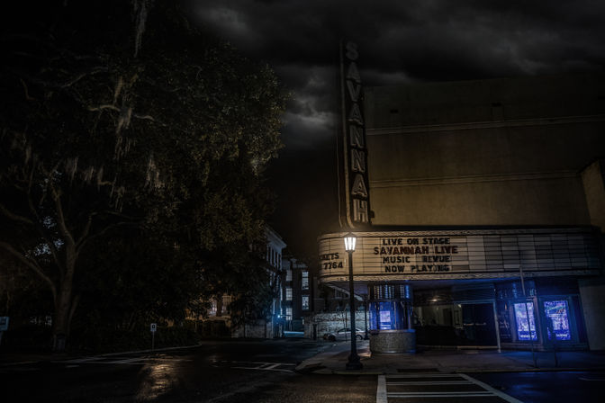 Wet downtown Savannah street at night featuring a lit vintage theater marquee and vertical sign, glowing lamppost, moss-draped oak trees, and stormy clouds.