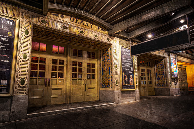 Ornate historic theater entrance at night with gold double doors, decorative tiled columns, illuminated marquee and poster displays along a covered downtown sidewalk.