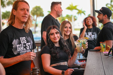 Smiling group of friends at a waterfront outdoor bar patio with palm trees, enjoying lime‑garnished cocktails.
